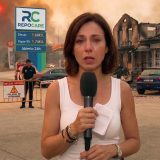 Una reportera con micrófono informa en directo desde una calle acordonada por la policía, con un edificio calcinado a un lado, una vecina de espaldas al otro lado de la valla y el frente del incendio al fondo