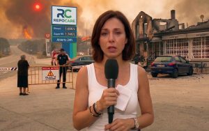 Una reportera con micrófono informa en directo desde una calle acordonada por la policía, con un edificio calcinado a un lado, una vecina de espaldas al otro lado de la valla y el frente del incendio al fondo